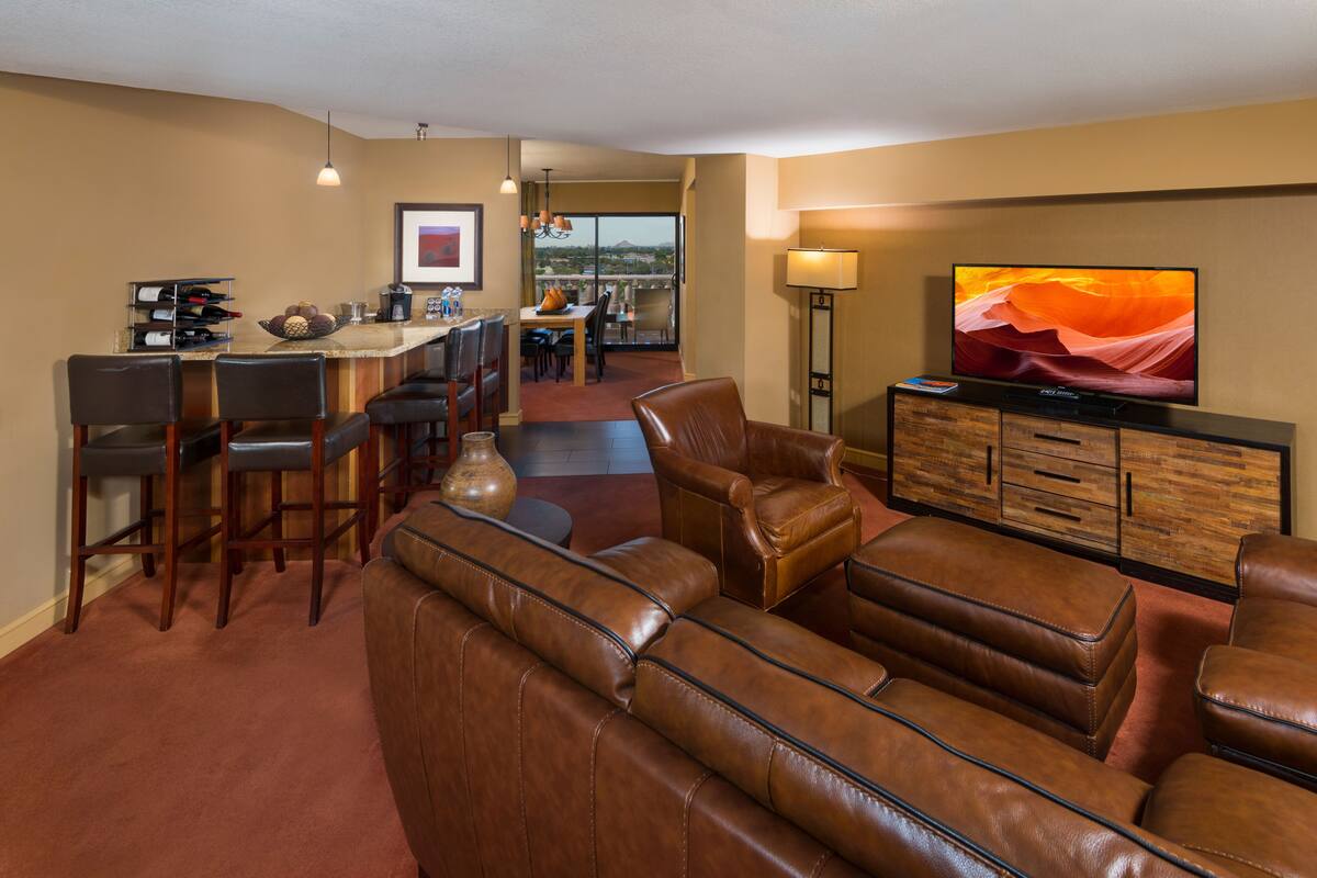 A modern living room with a leather sofa, a dining area with a bar, and a large TV on the wall.