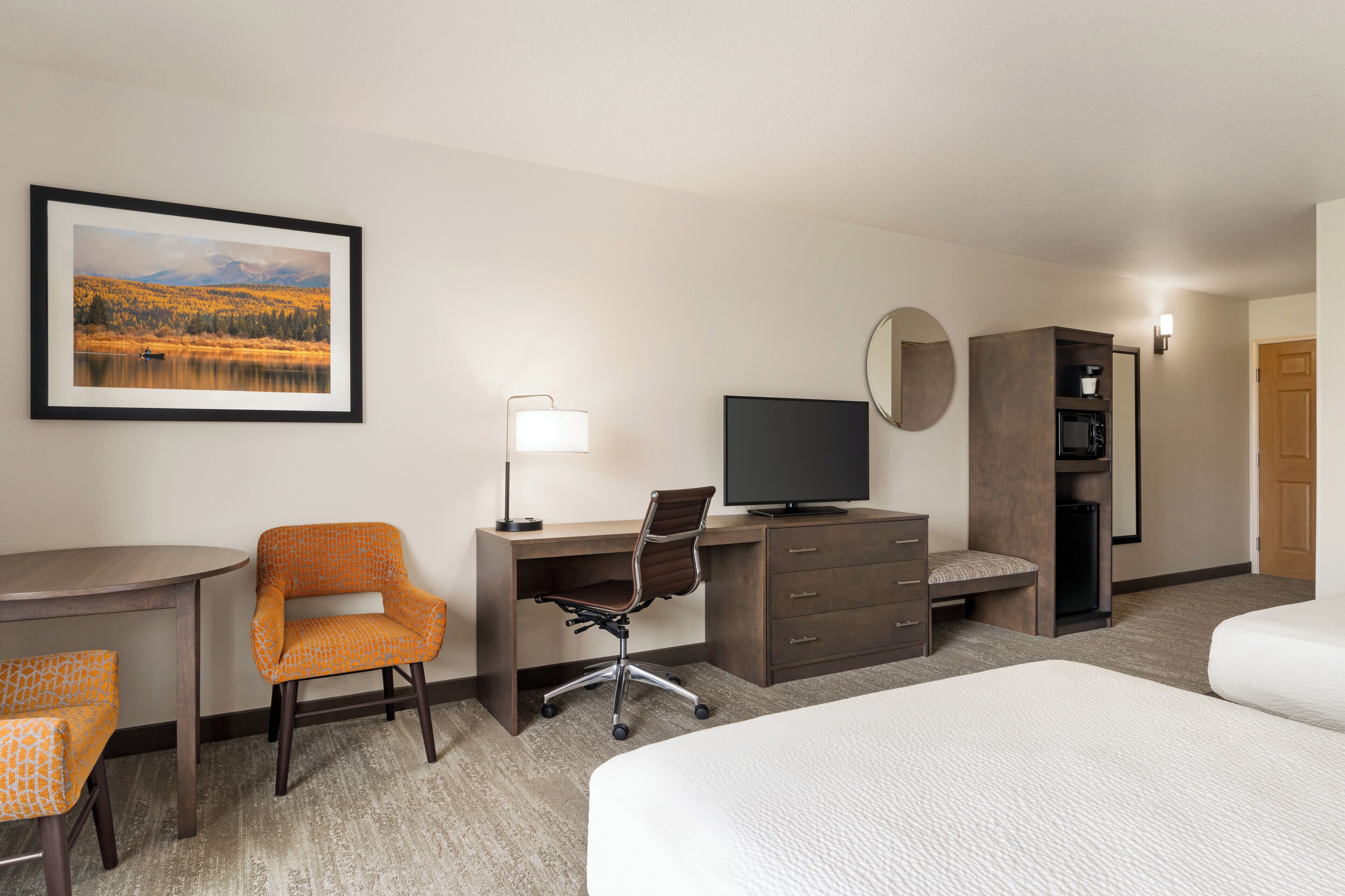 A hotel room with a bed, desk, chair, TV, dresser, and a framed landscape picture on the wall.