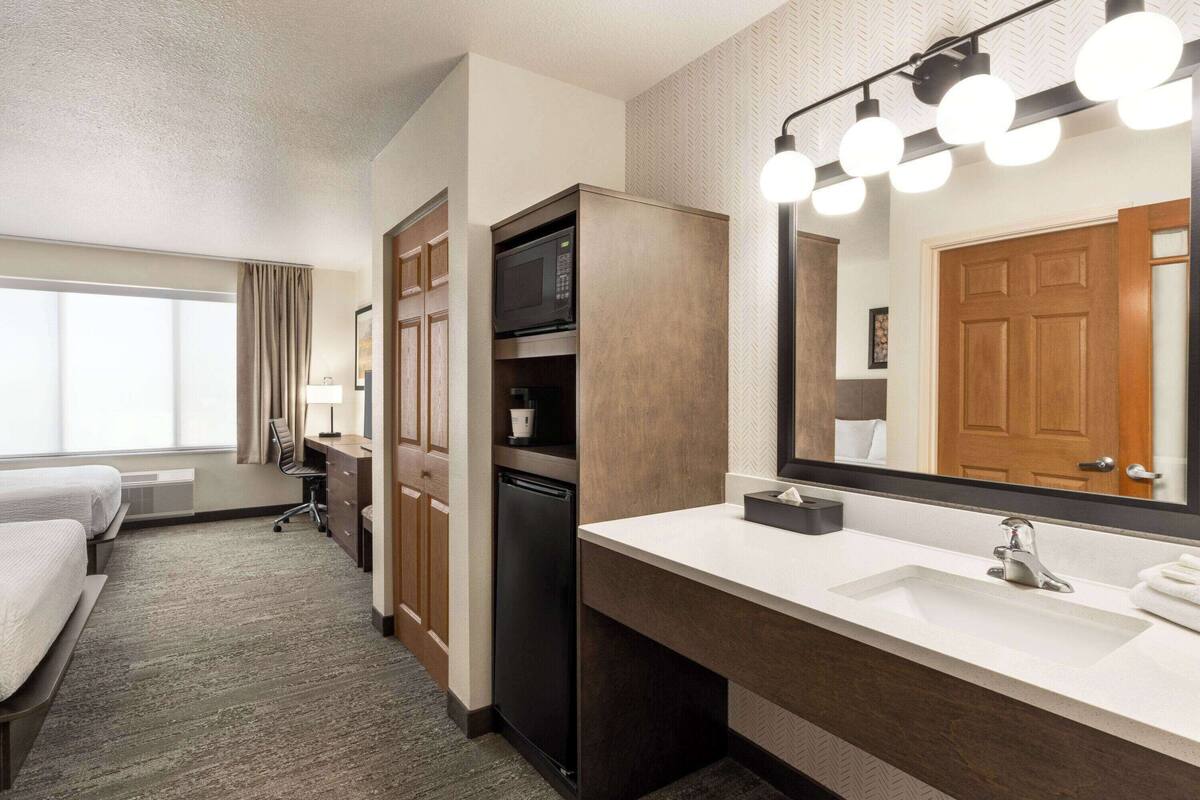 Ett hotellrum med en stor säng, ett skrivbord, en mikrovågsugn, ett kylskåp, ett handfat och en spegel med belysning.