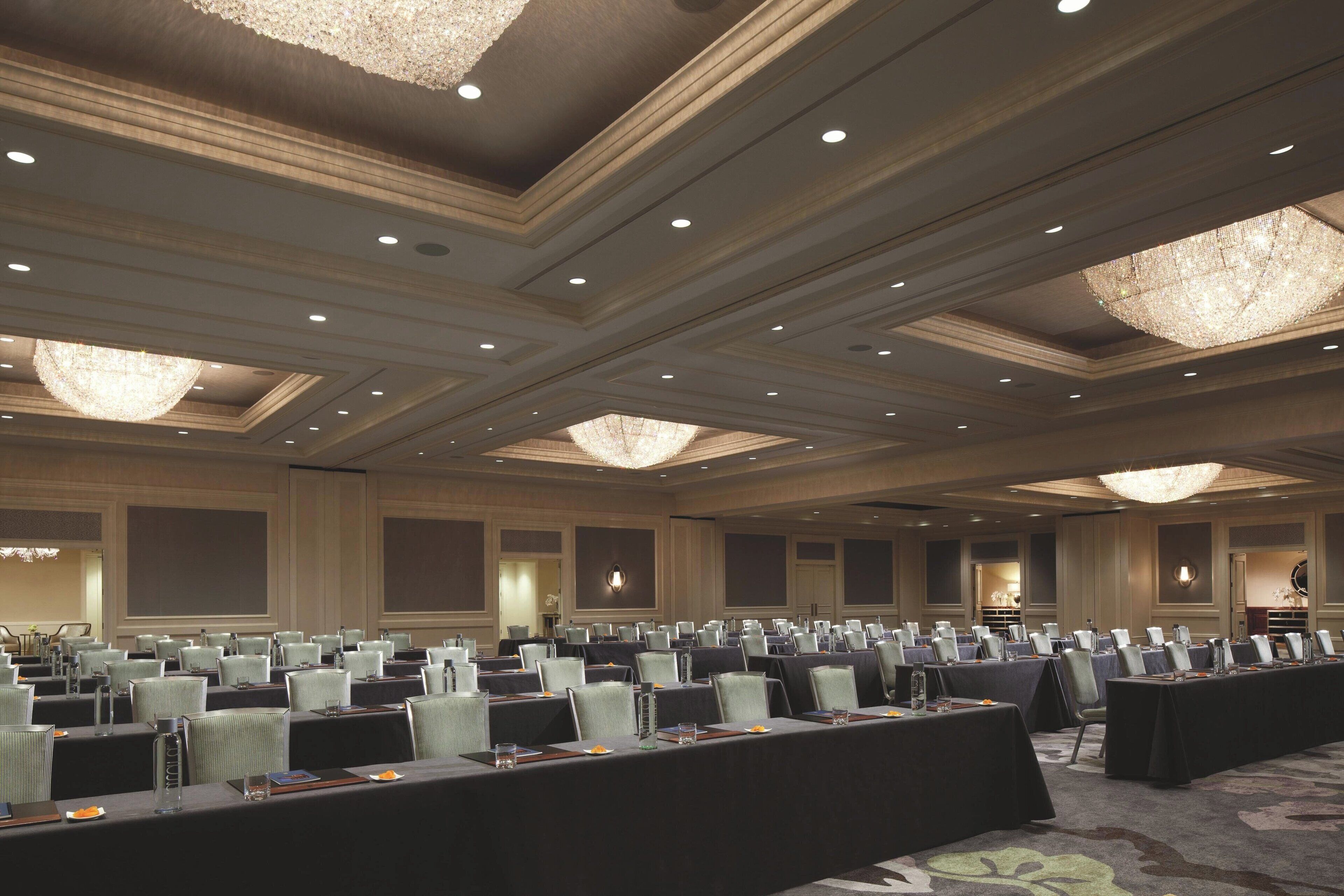 A conference room with rows of tables and chairs, large chandeliers, and a carpeted floor.