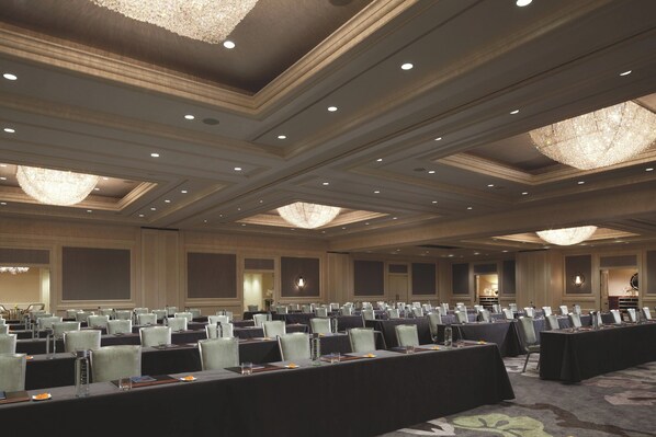 A conference room with rows of tables and chairs, large chandeliers, and a carpeted floor.