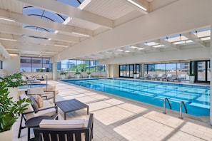 A modern indoor swimming pool with clear blue water, surrounded by lounge chairs and tables, and large windows offering views of the city.