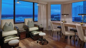 Camera d'hotel moderna con un'ampia finestra che offre vista sulla città, un'area salotto accogliente con poltrona massaggiante, un tavolo da pranzo con sedie e un piccolo tavolino con lampada.