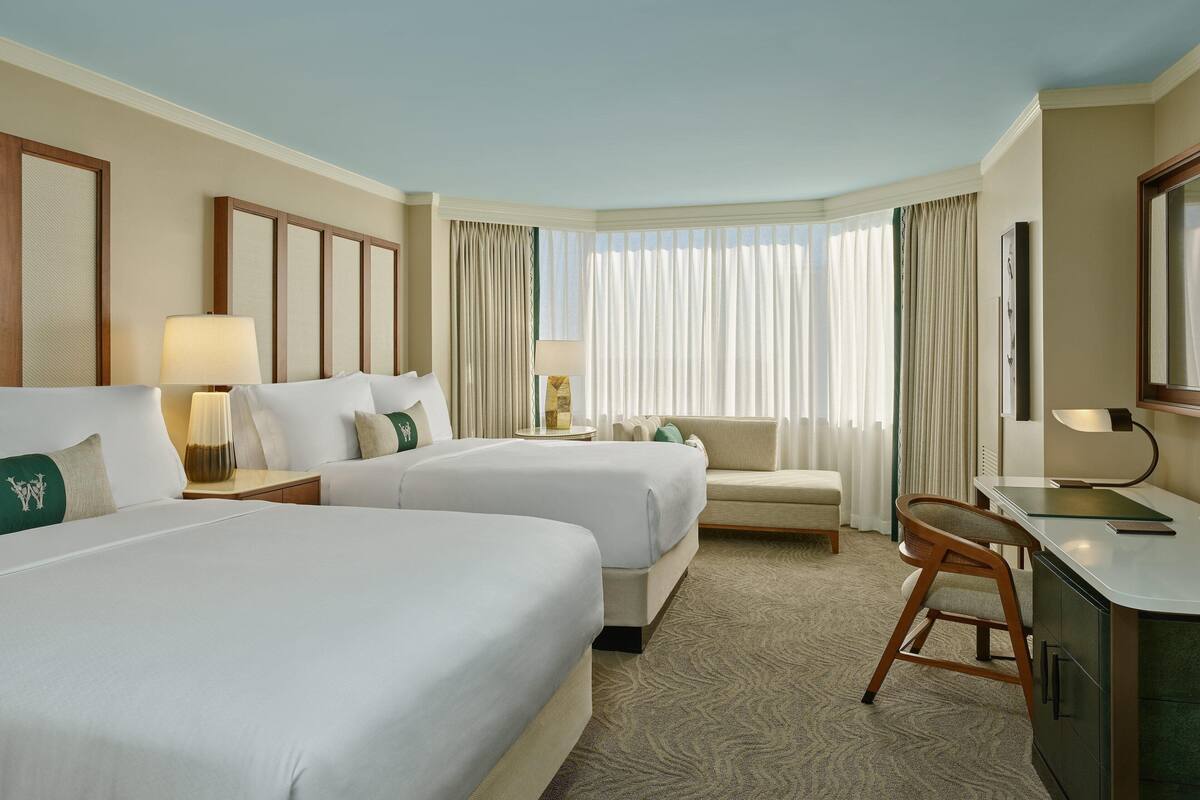 A hotel room with two beds, a sofa, a desk, and a large window with curtains.