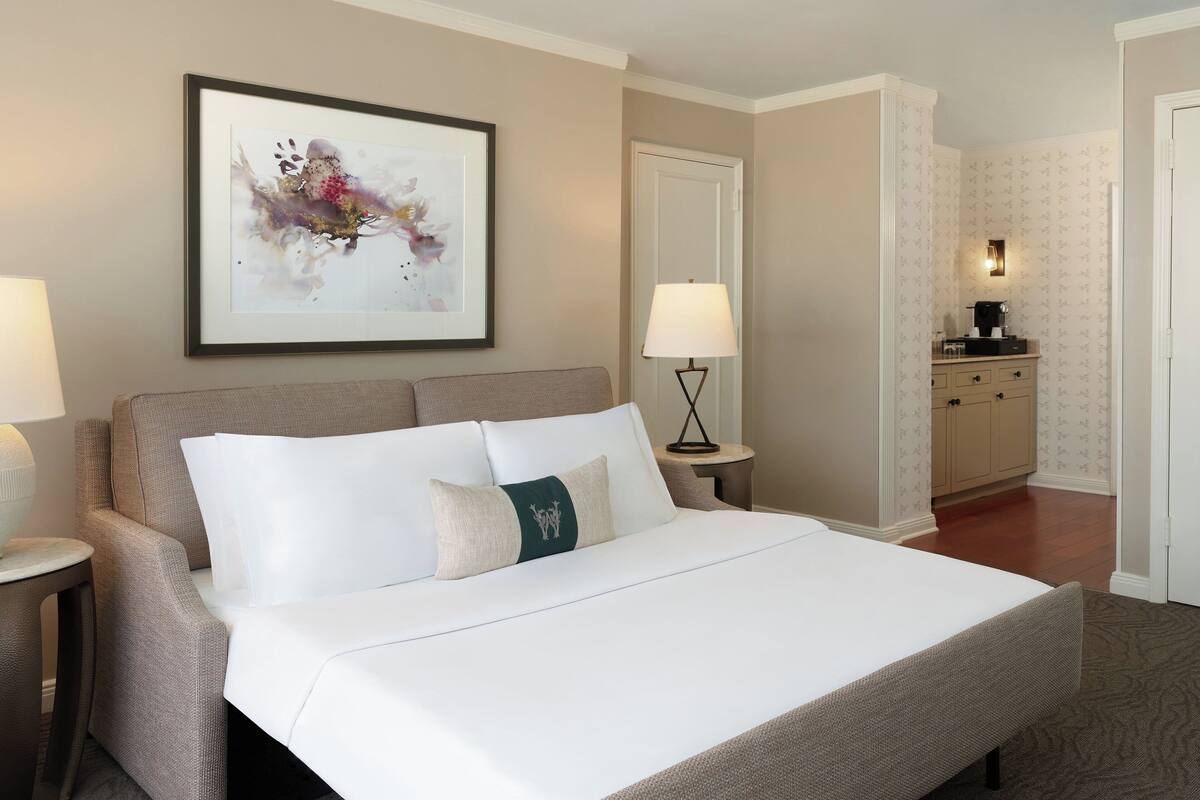 A neatly made bed with white linens and a green accent pillow, a framed abstract painting on the wall, a bedside lamp, and a small table with a lamp.