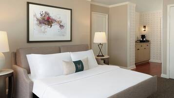 A neatly made bed with white linens and a green accent pillow, a framed abstract painting on the wall, a bedside lamp, and a small table with a lamp.