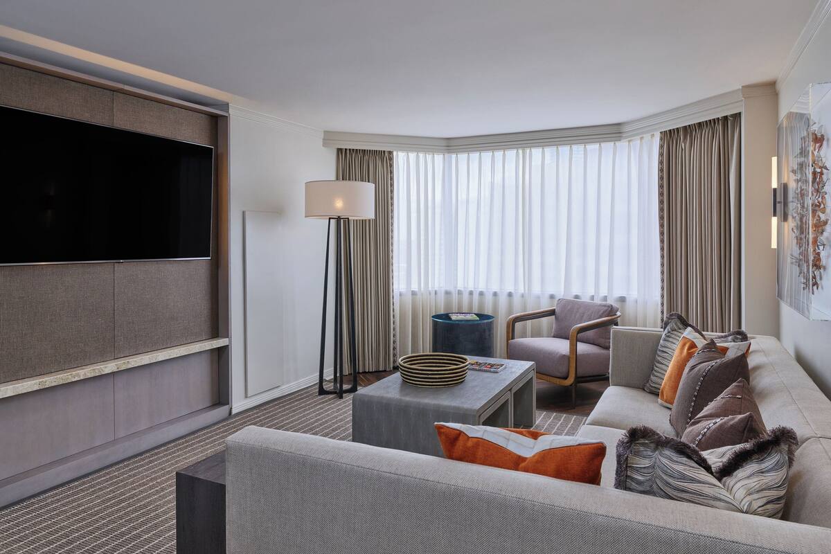 A modern hotel room with a large flat-screen TV, a sofa, a chair, a coffee table, and a floor lamp.