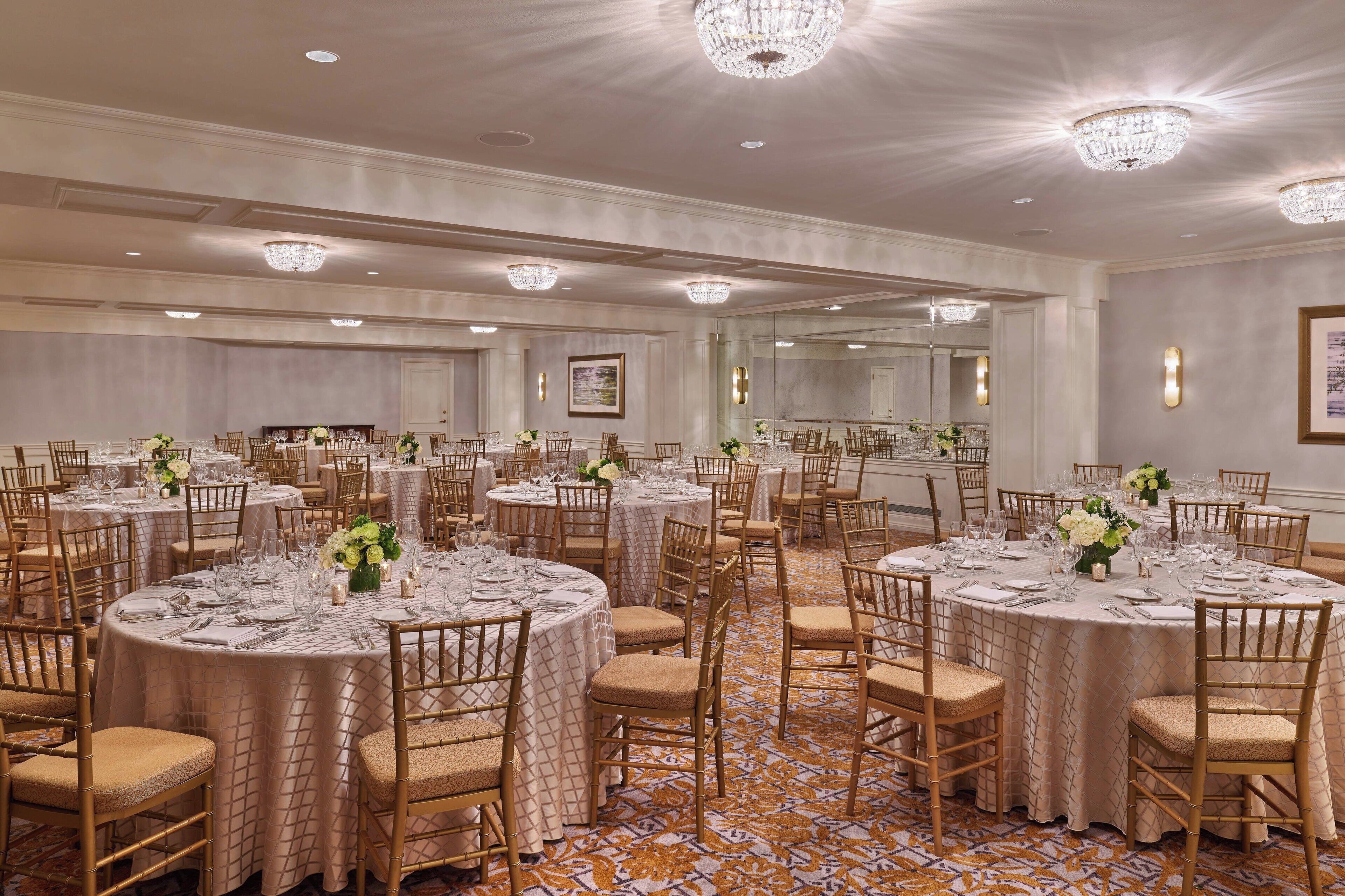 A banquet hall with round tables, gold chairs, and chandeliers.