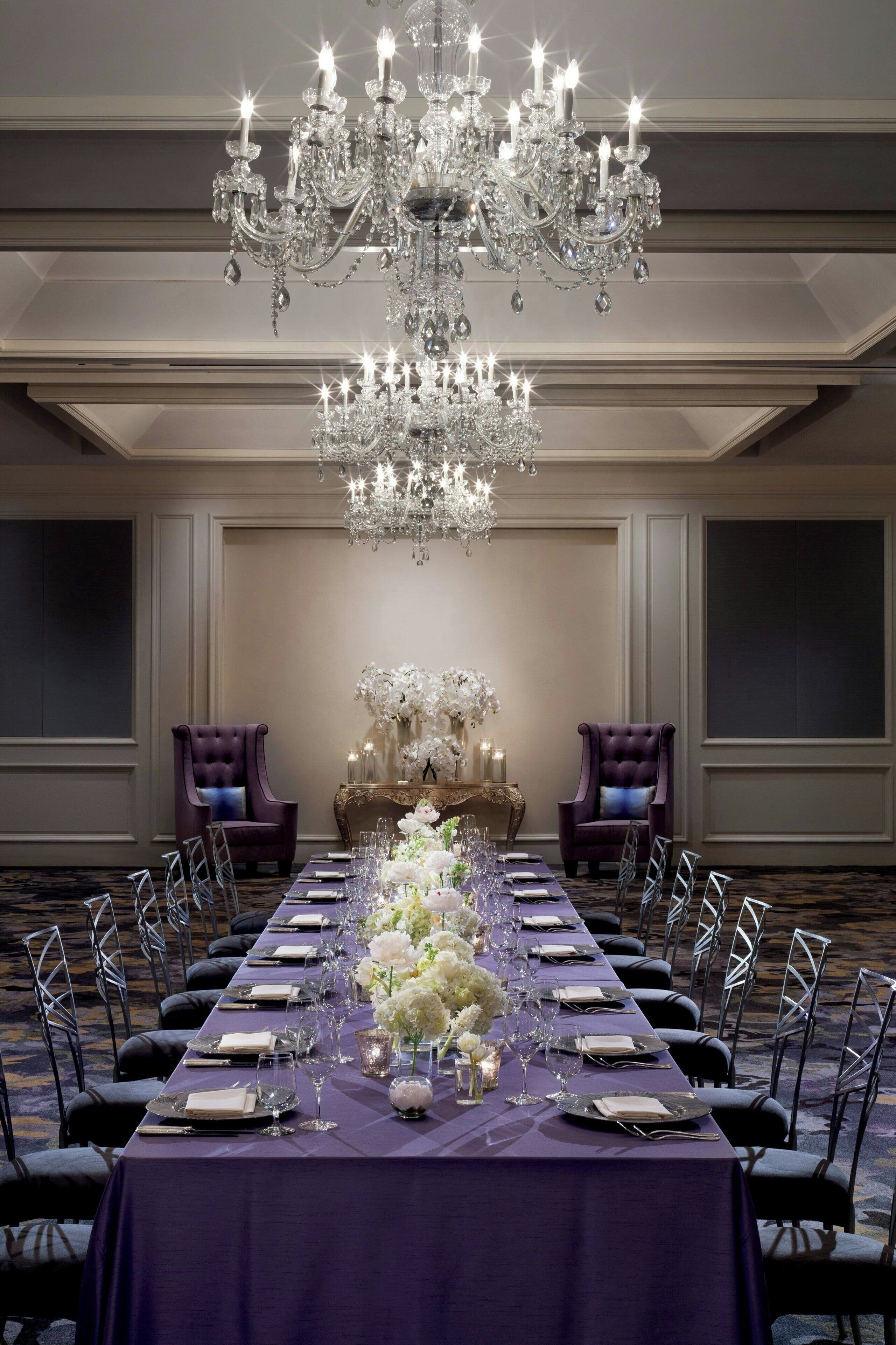 A formal dining room with a long table set for a meal, flanked by two purple armchairs, a chandelier, and a floral centerpiece.