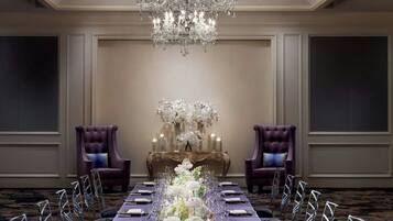 A formal dining room with a long table set for a meal, flanked by two purple armchairs, a chandelier, and a floral centerpiece.