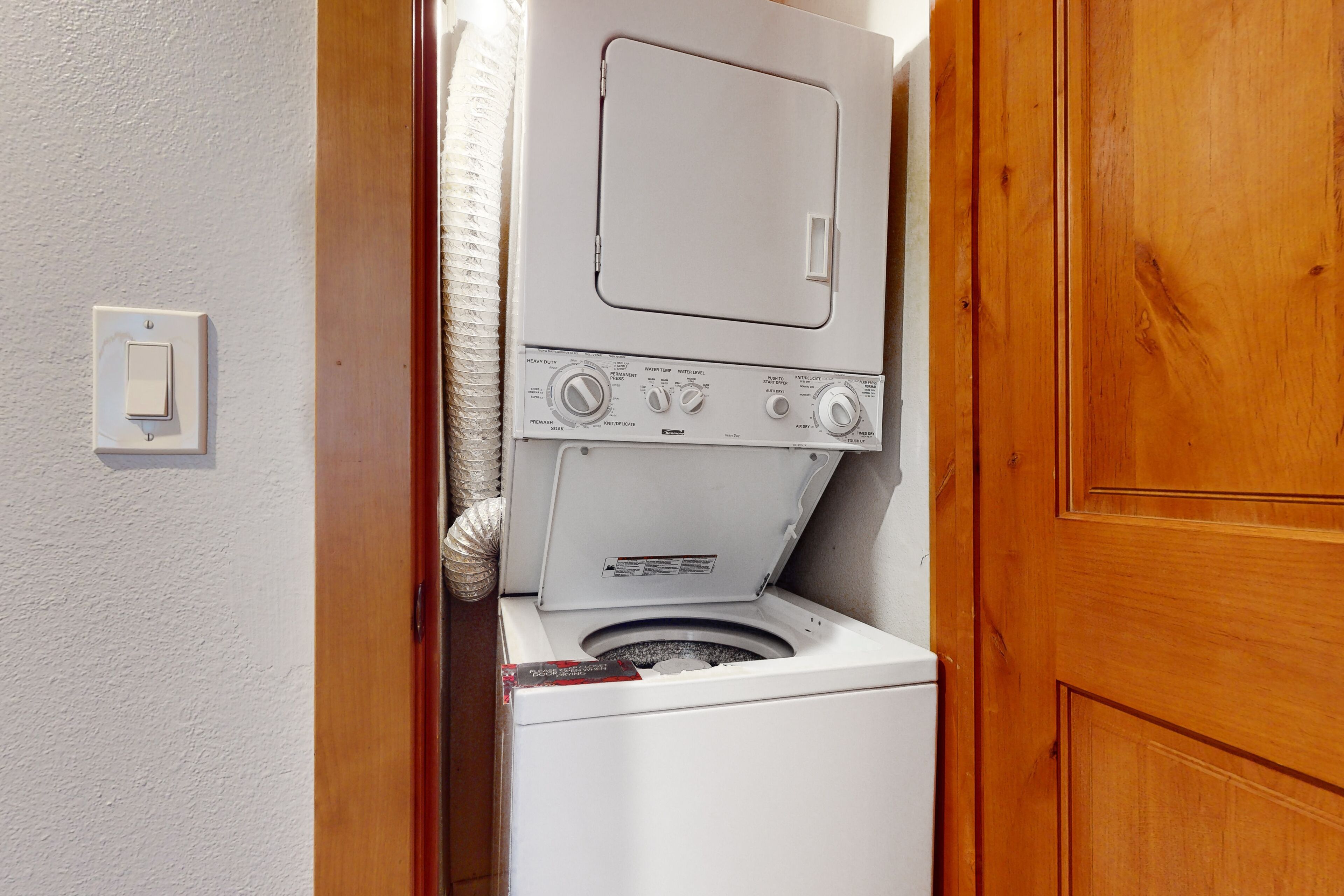 A compact laundry room with a stacked washer and dryer, a wooden door, and a light switch on the wall.