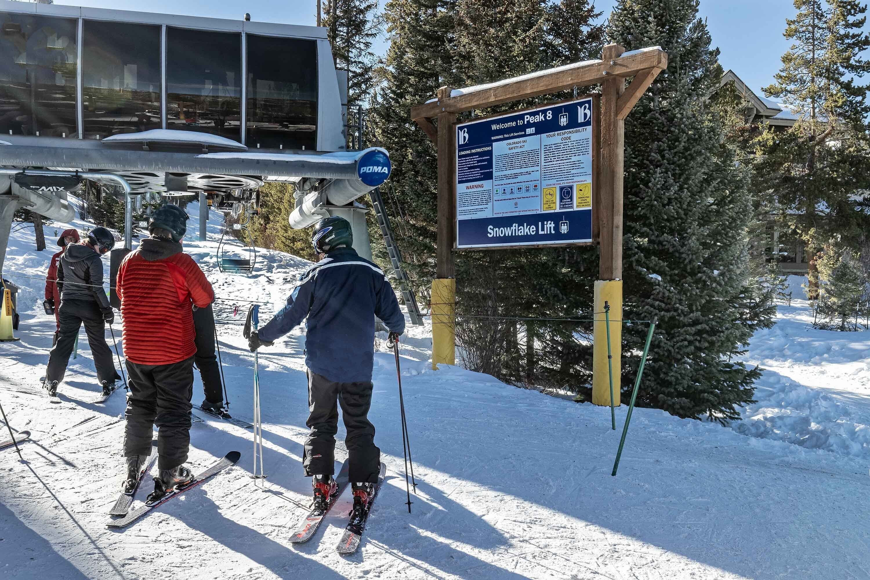 Skiers at a lift station with a sign indicating 'Welcome to Peak 8' and 'Snowflake Lift'.