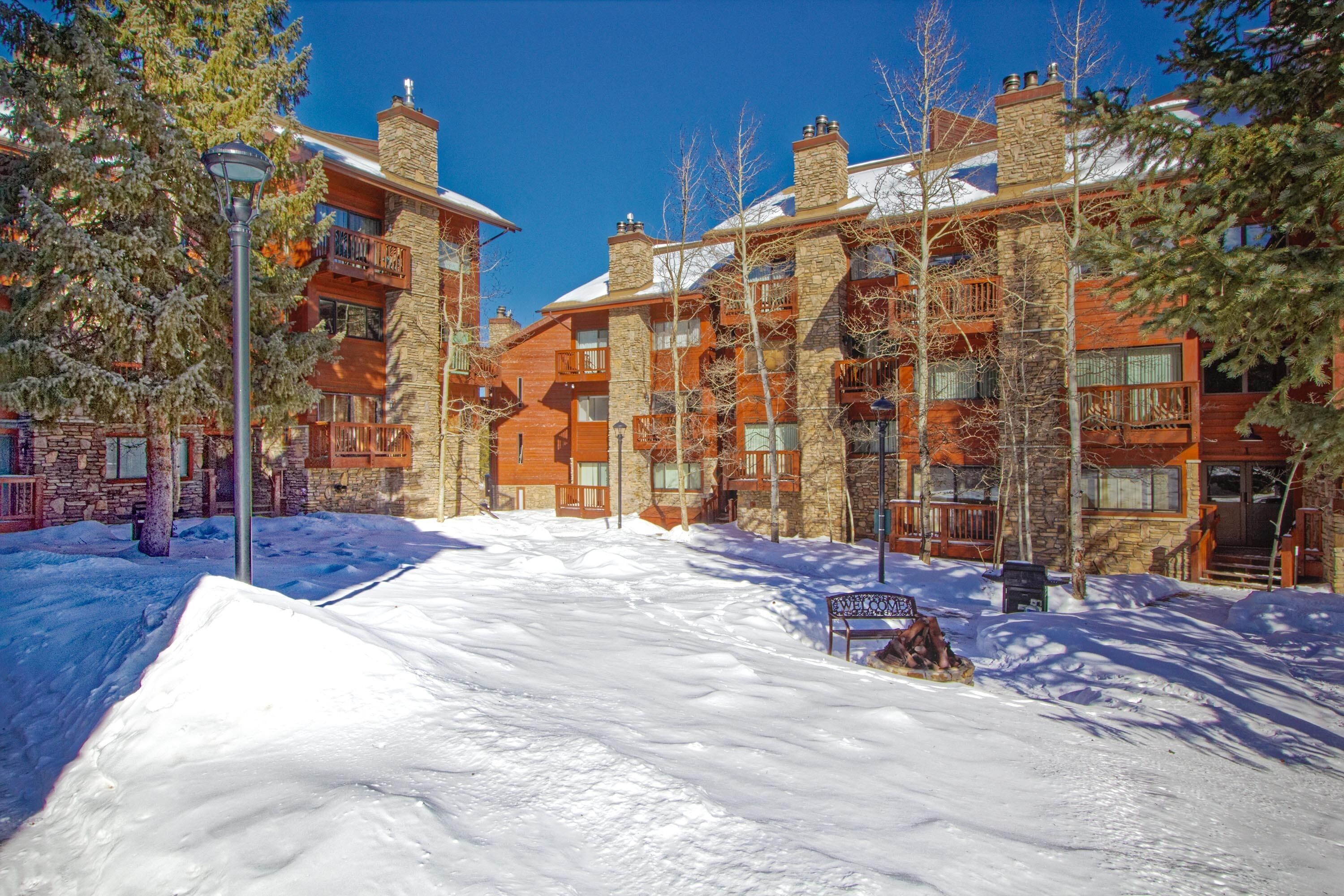 A snow-covered courtyard with a wooden bench, stone buildings with balconies, and a clear blue sky.