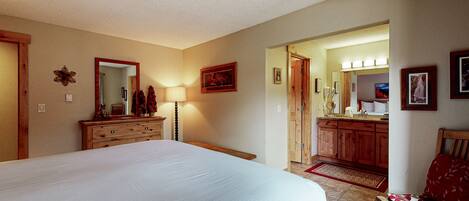 Une chambre avec un grand lit, une commode en bois et un miroir. Une porte donne accès à une autre pièce où se trouvent une coiffeuse et un miroir.