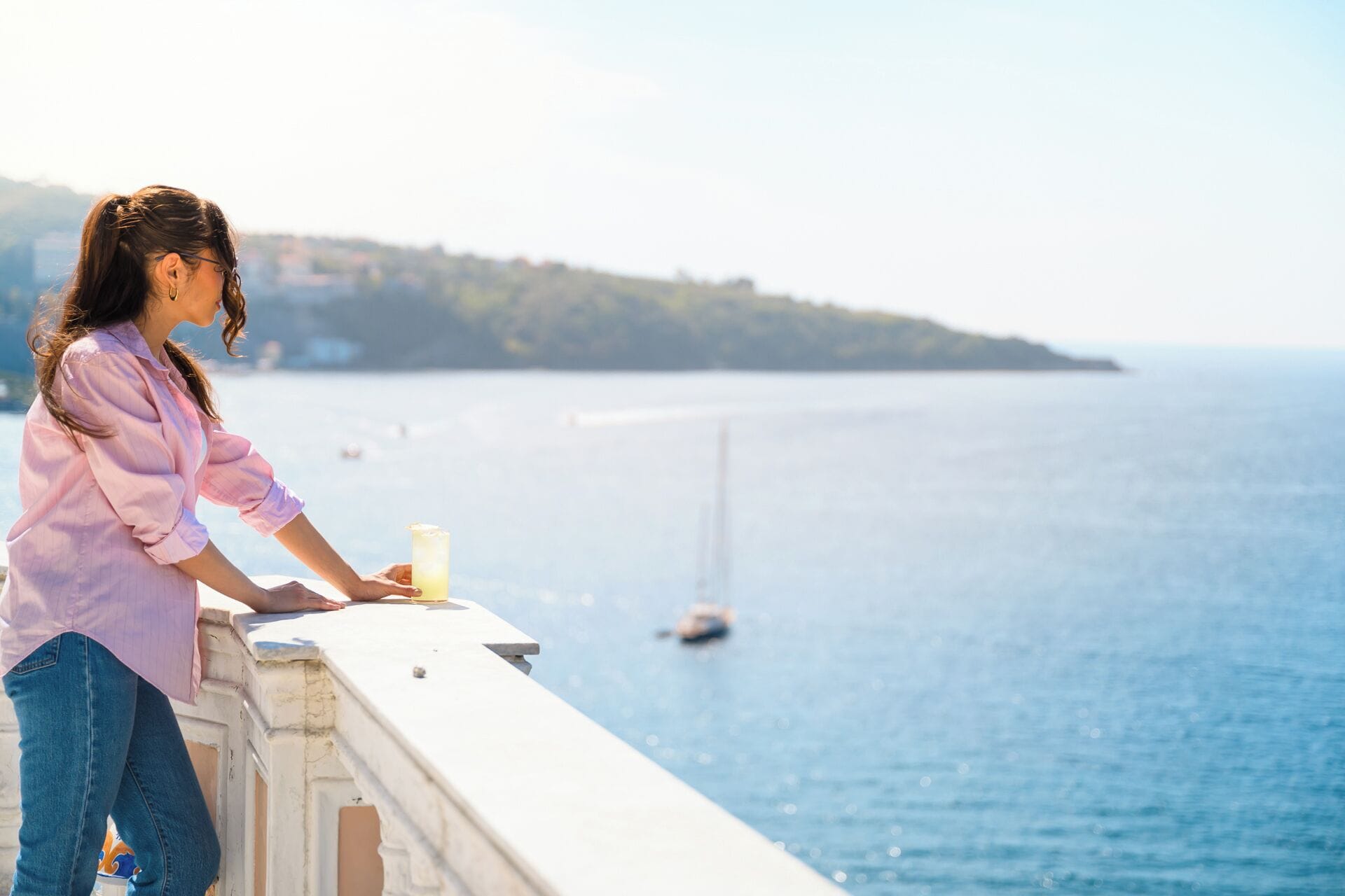 A person leaning on a balcony overlooking a body of water with boats and a distant landmass.