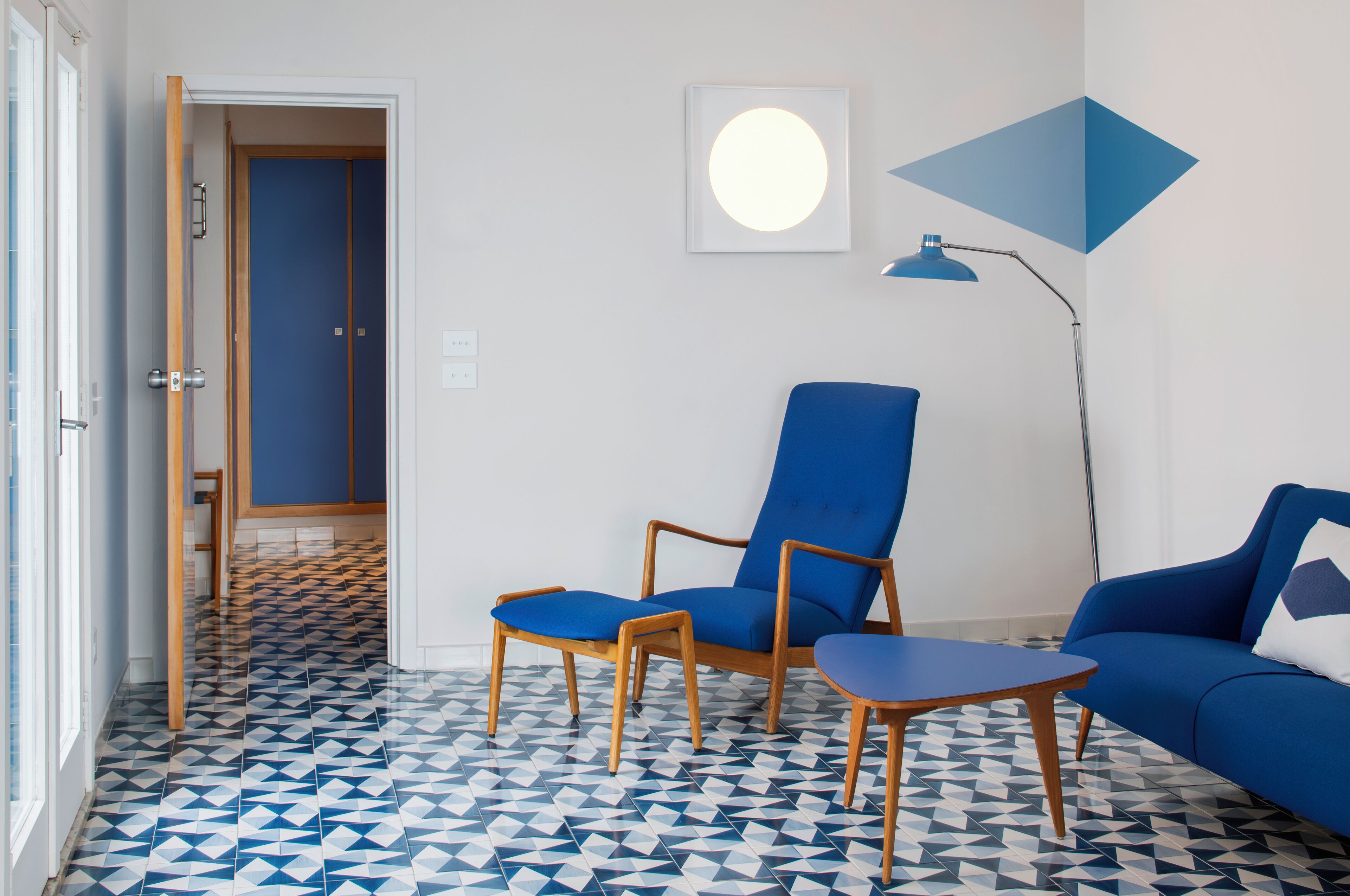 Quarto com piso de padrão geométrico, um sofá azul, uma poltrona azul e uma pequena mesa.