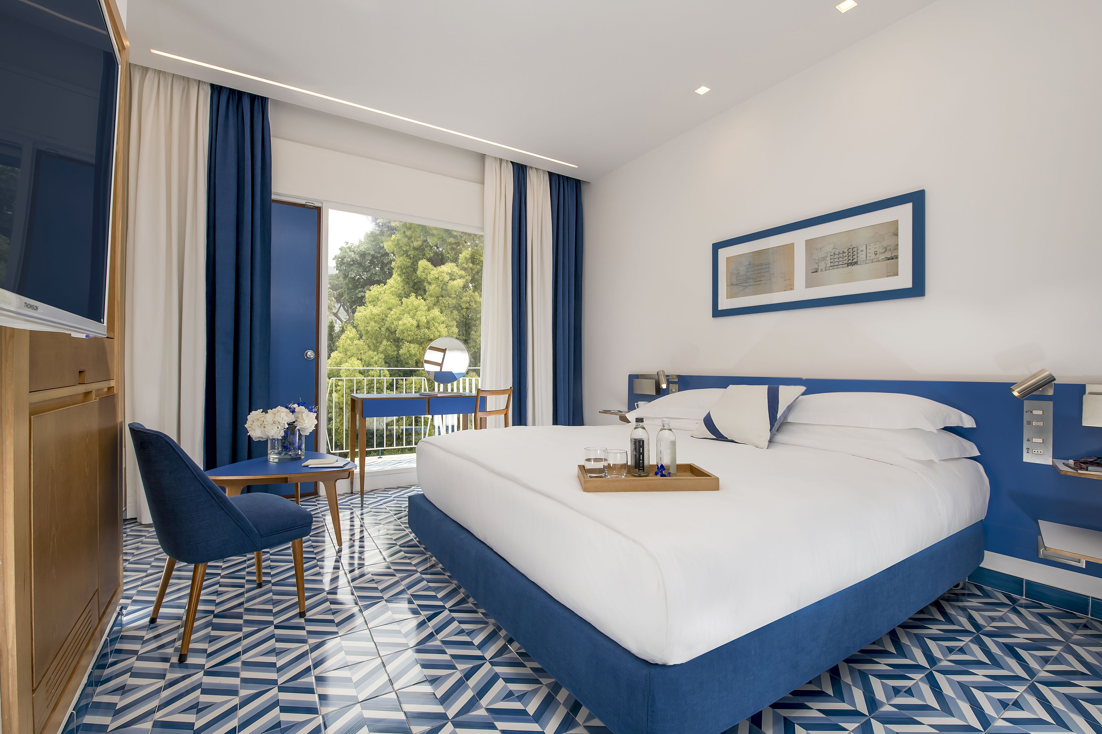 A hotel room with a large bed, a small table with a vase of flowers, a chair, and a balcony with a view of trees.
