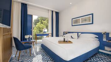 A hotel room with a large bed, a small table with a vase of flowers, a chair, and a balcony with a view of trees.