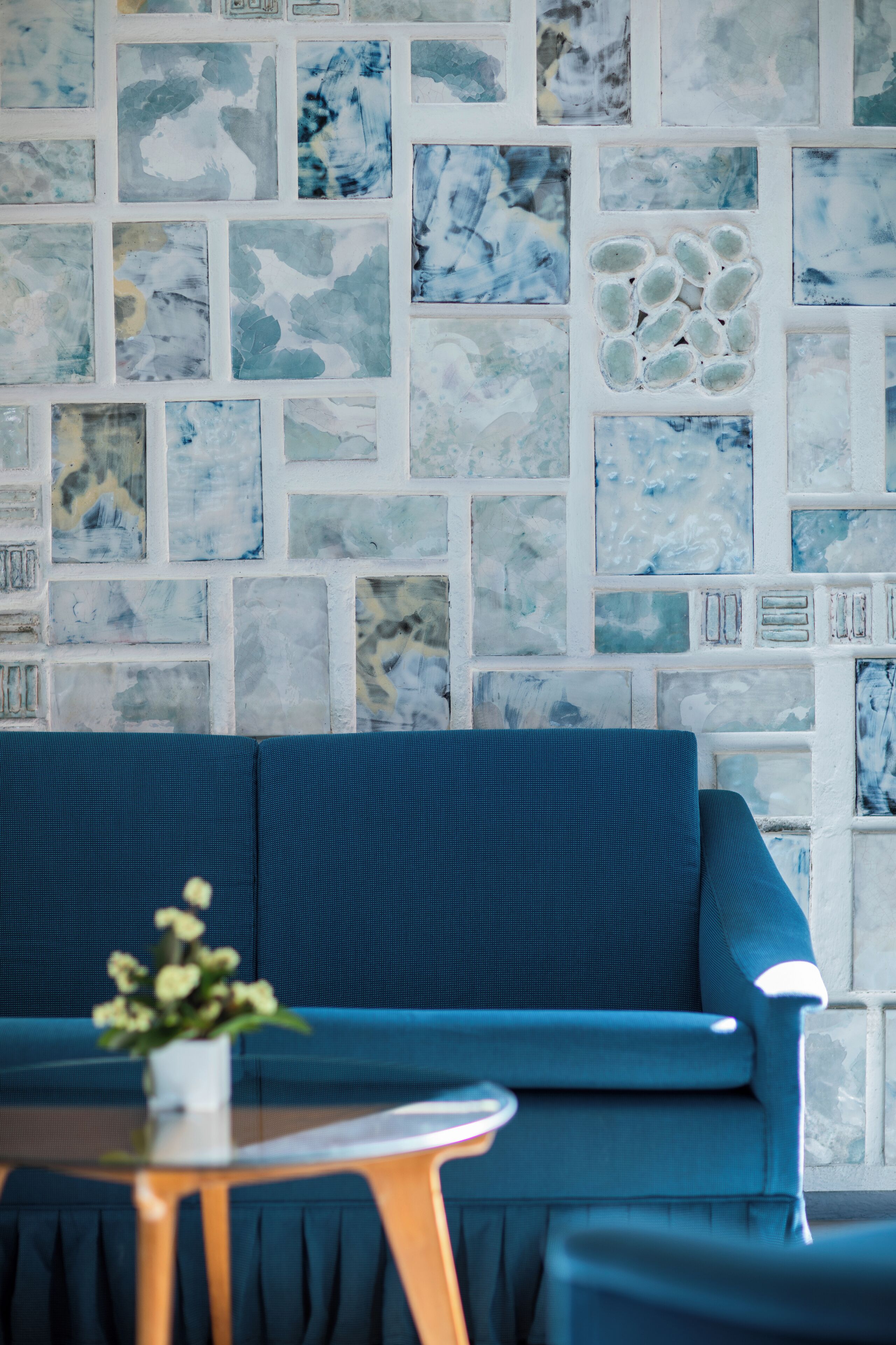 Un sofá azul con una mesita y una planta frente a una pared con diseño de mosaico.