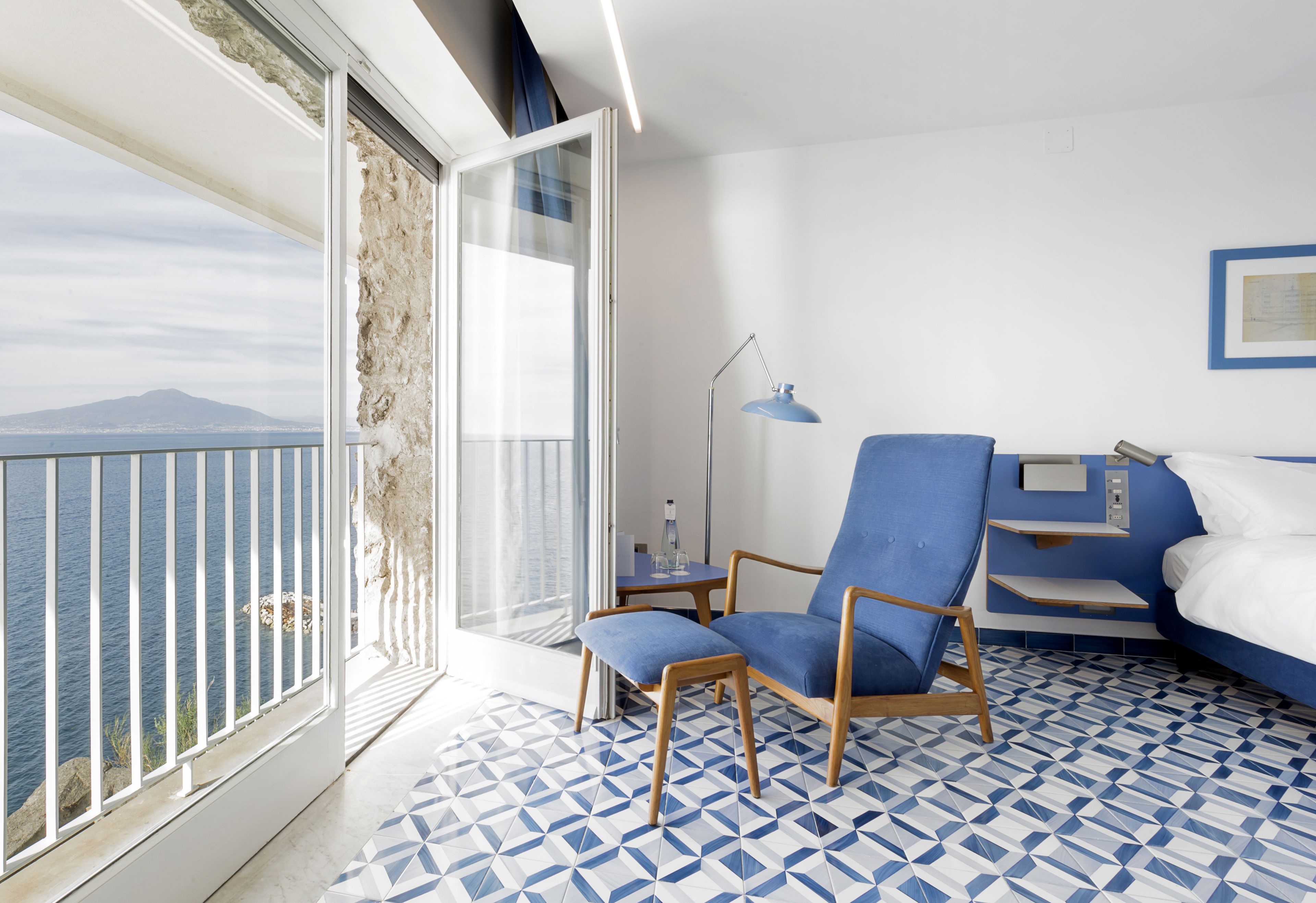 Une pièce avec une grande fenêtre, un fauteuil bleu, un lit et un sol à motifs.