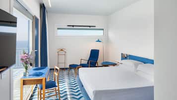 A hotel room with a bed, a desk with a chair, a blue armchair, a hanging light, and a window with a view.