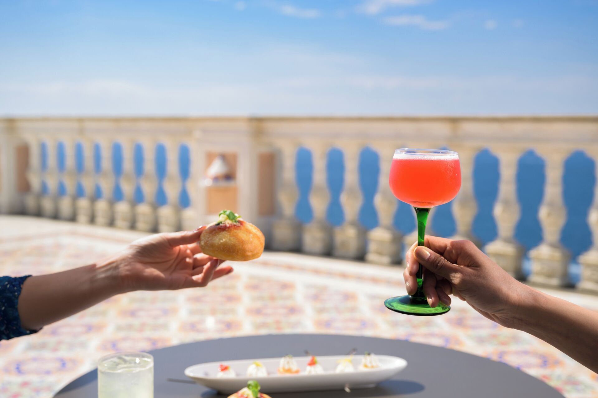 Una terraza con una mesa preparada para comer, una bebida en un vaso y un pastel en la mano de alguien.