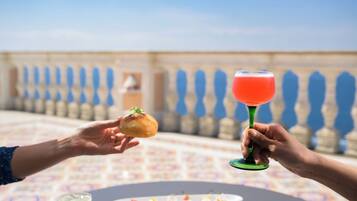 Una terraza con una mesa preparada para comer, una bebida en un vaso y un pastel en la mano de alguien.