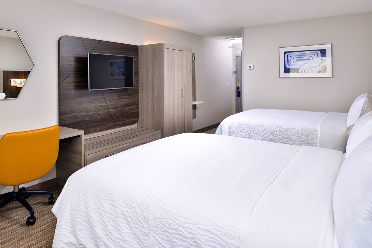 A hotel room with two beds, a desk with a chair, a TV mounted on a wooden panel, and a painting on the wall.