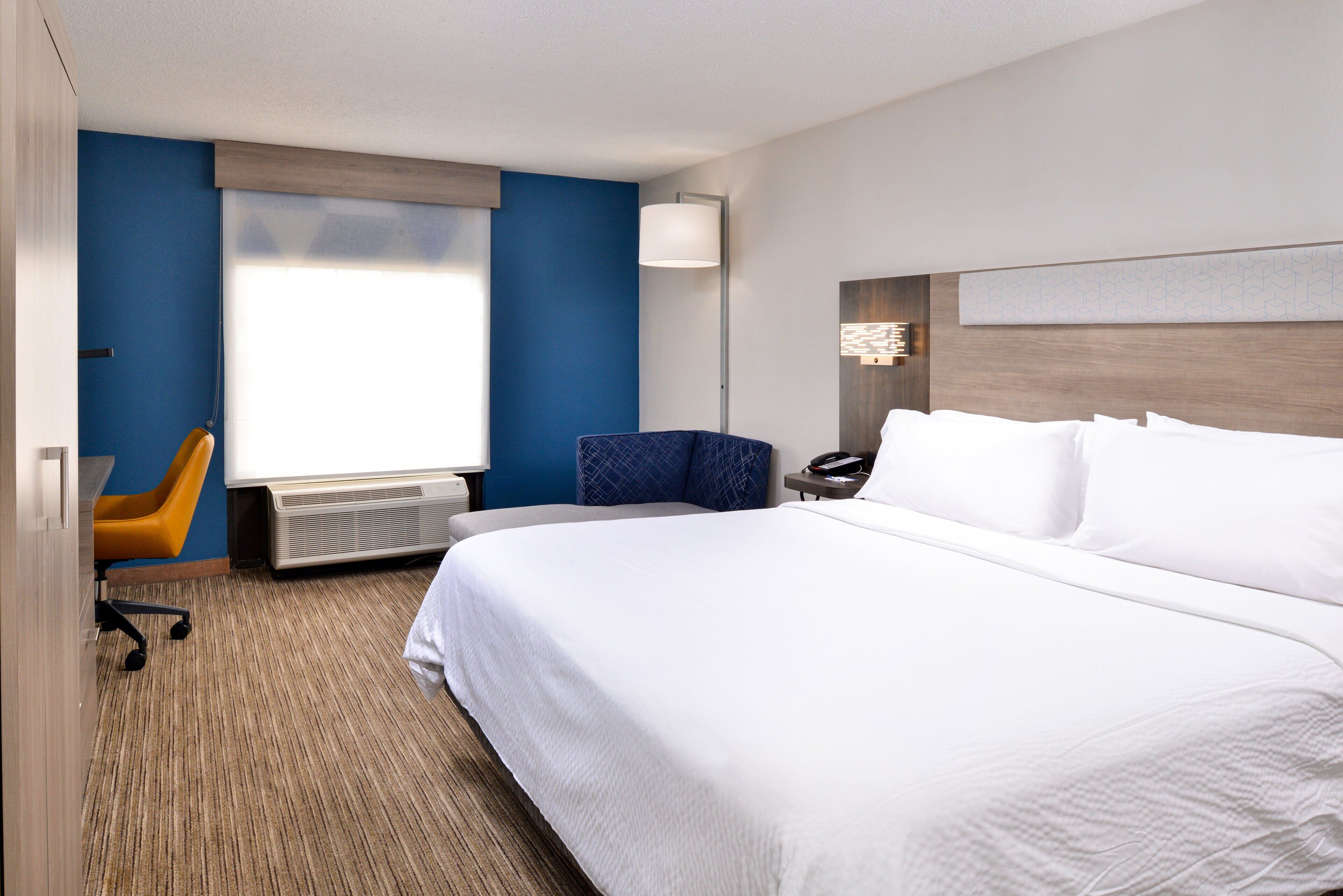 Habitación de hotel con una cama grande, un escritorio con silla, una ventana con persianas y una lámpara.