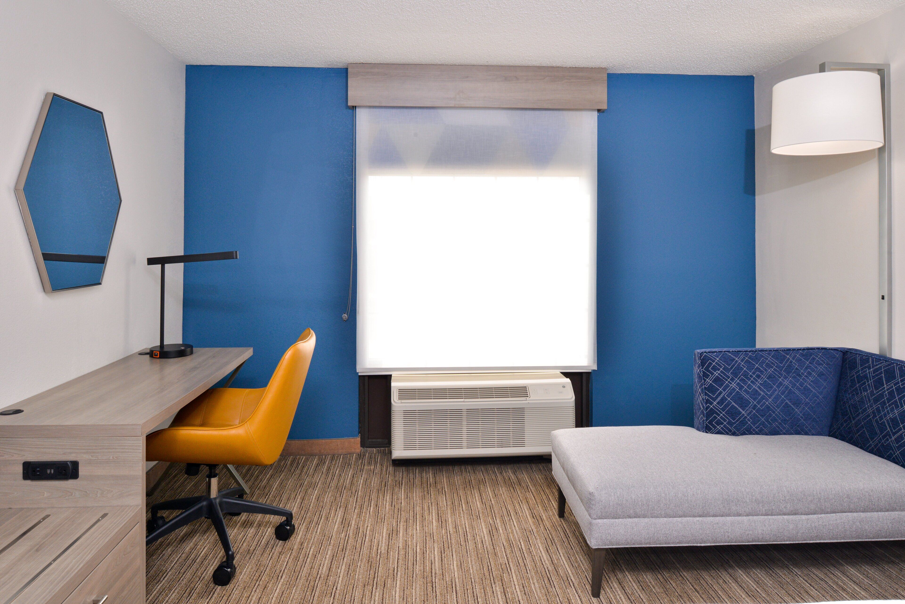 A hotel room with a desk, chair, and couch, a window with blinds, a standing lamp, and a wall-mounted air conditioner.