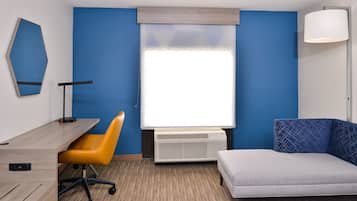 A hotel room with a desk, chair, and couch, a window with blinds, a standing lamp, and a wall-mounted air conditioner.