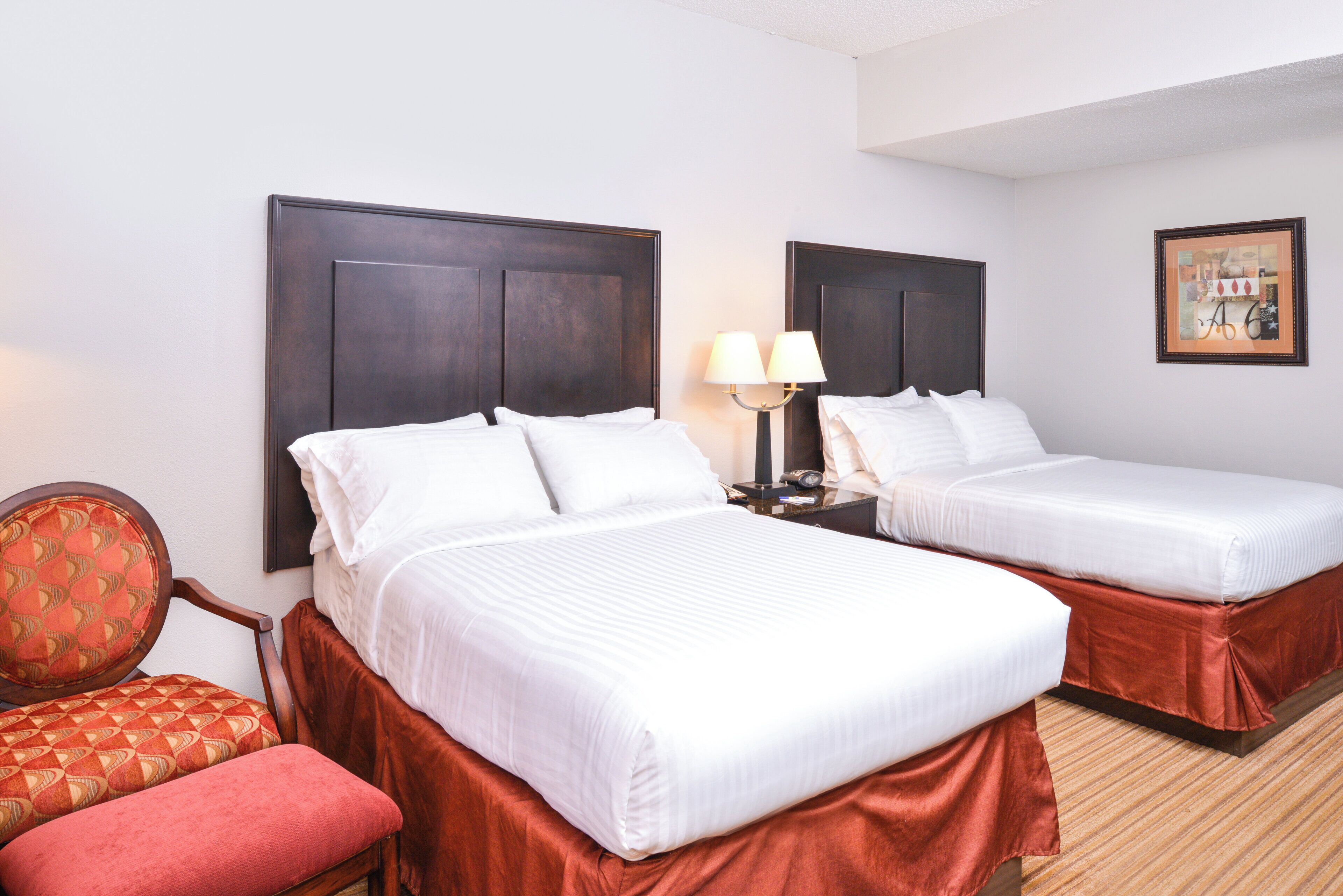 A hotel room with two beds, a nightstand with a lamp, a chair, and a framed picture on the wall.