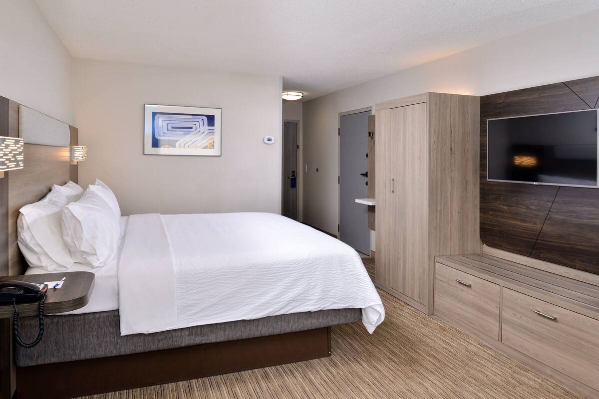 A hotel room with a bed, bedside table, lamp, TV, wardrobe, and a painting on the wall.
