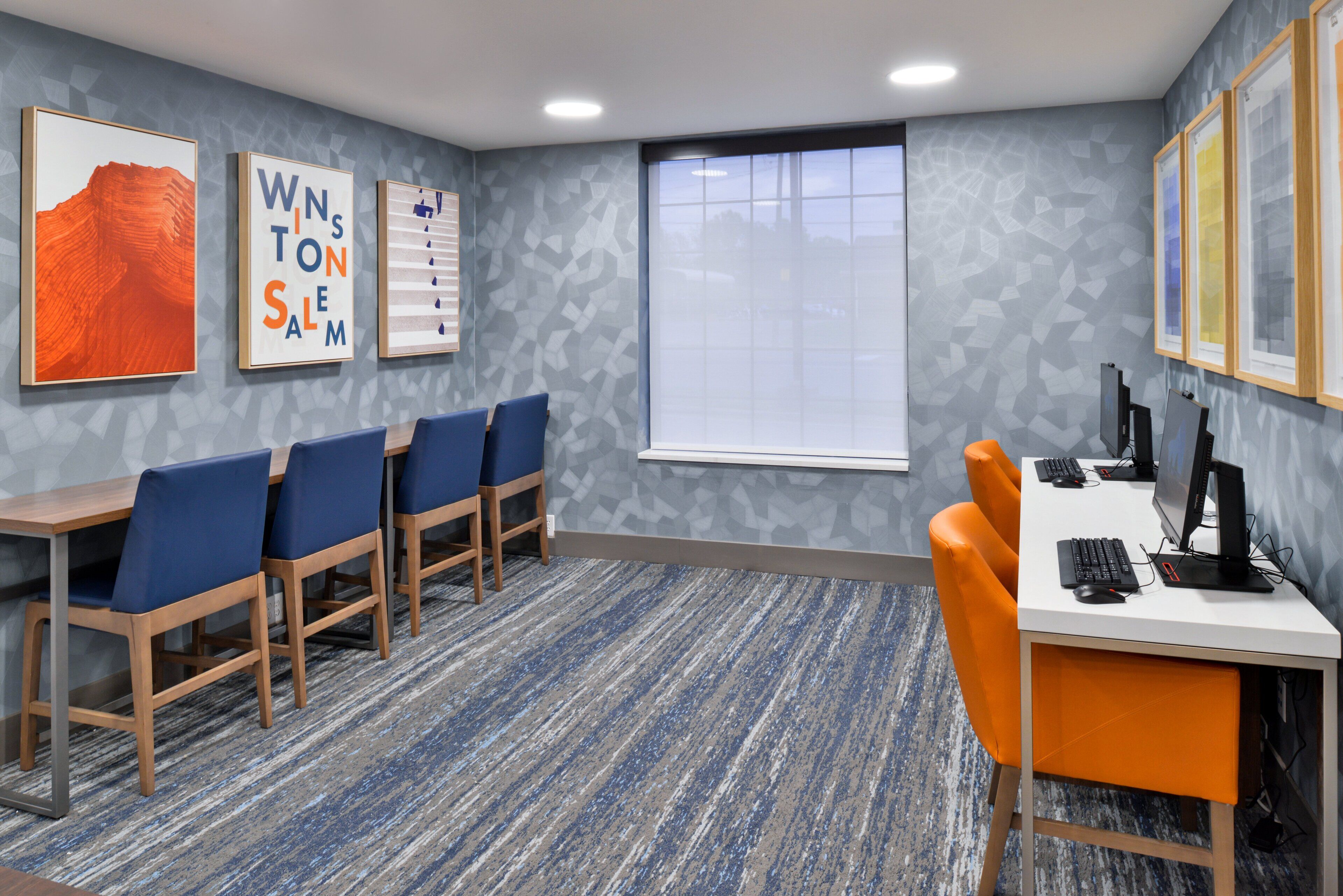 A conference room with a long table, blue and orange chairs, a white desk with a computer, and framed artwork on the walls.