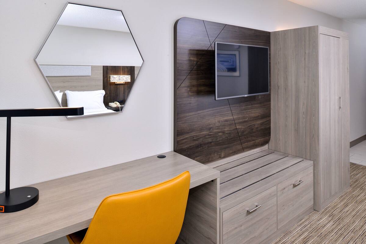 A hotel room with a wooden desk, a yellow chair, a large mirror, a flat-screen TV, and a wardrobe.