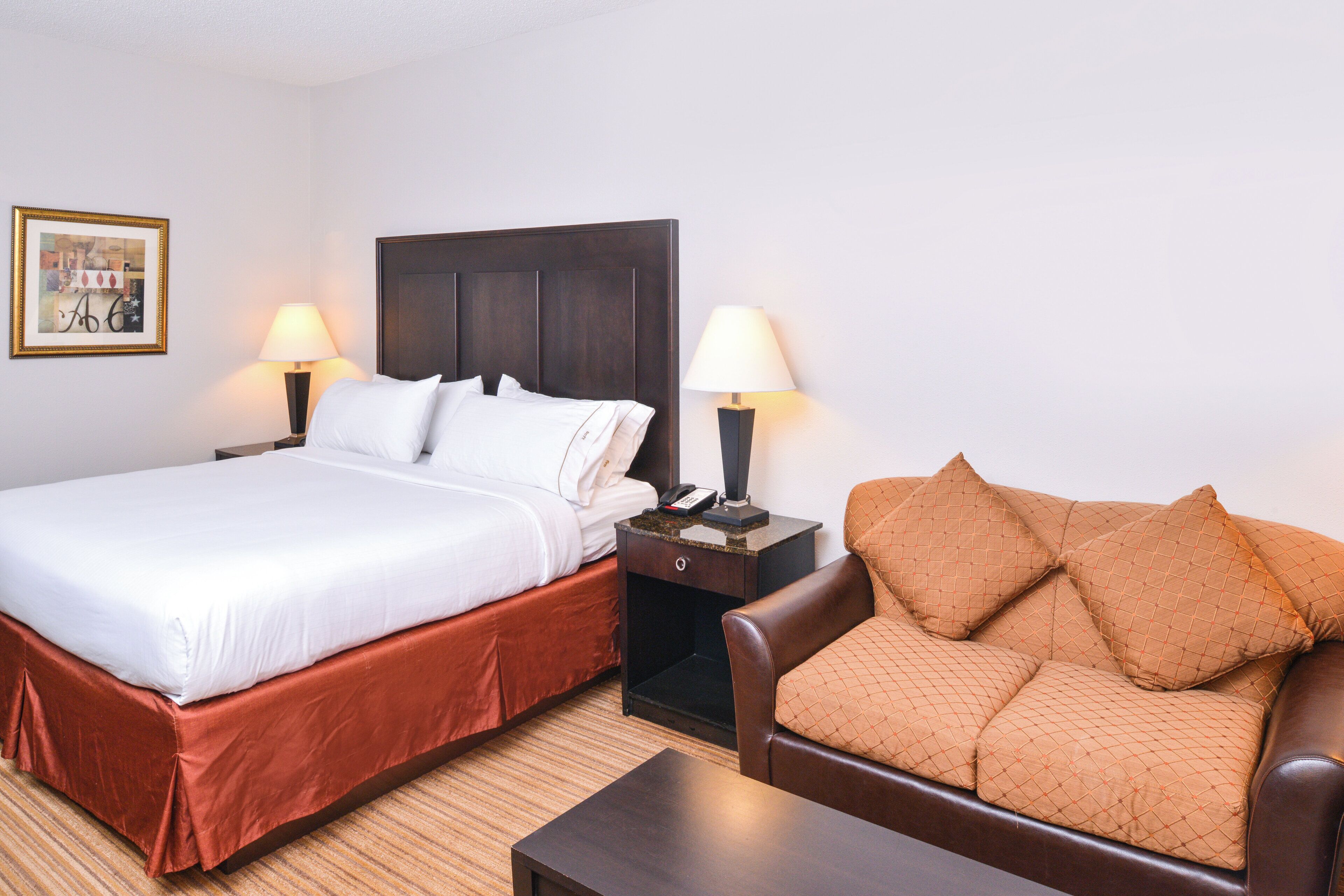Habitación de hotel con cama, dos lámparas de noche, un sofá de cuero marrón con cojines y una mesita.