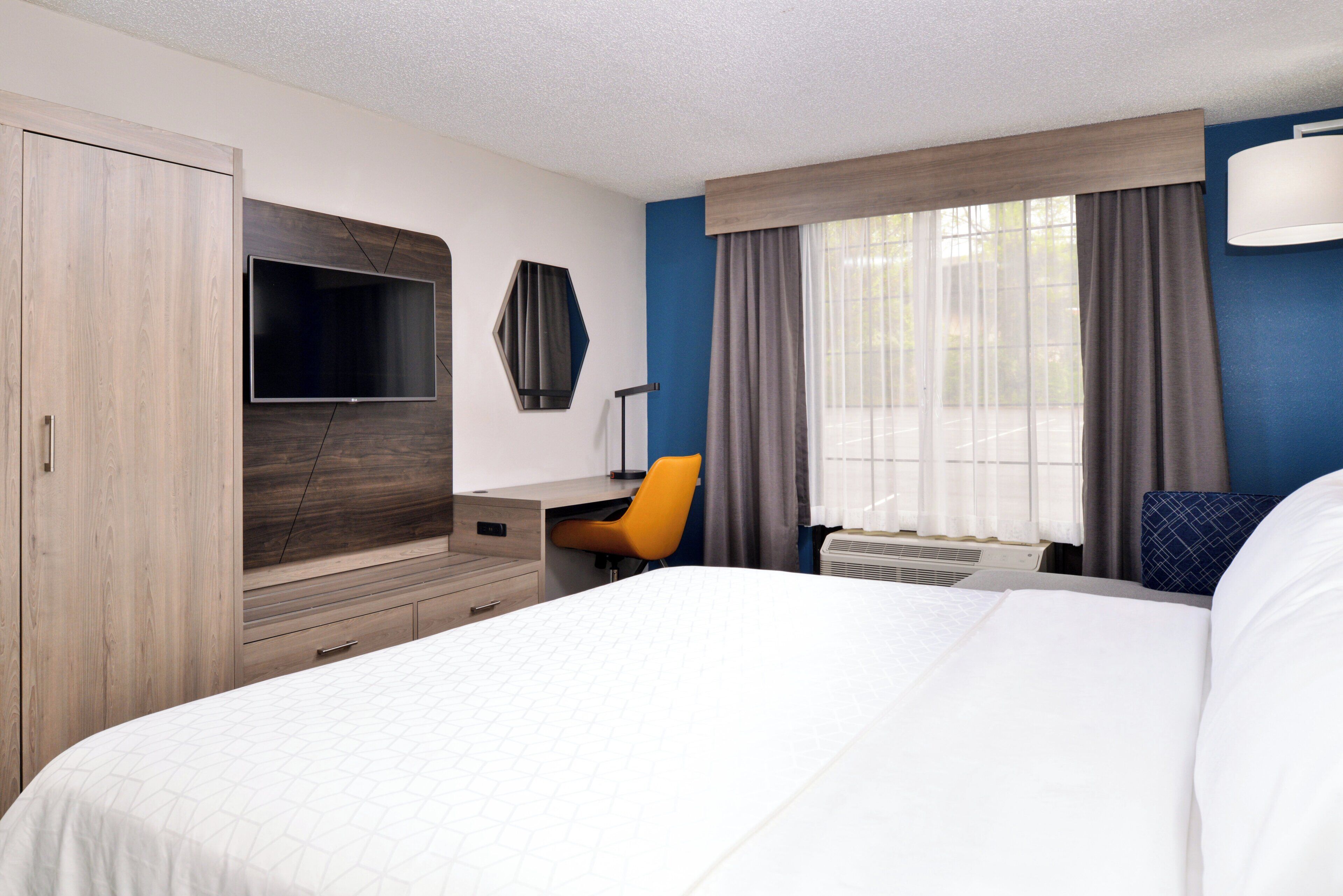 Una habitación de hotel con cama, escritorio, silla, televisión y ventana con cortinas.