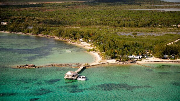 An aerial view of a coastal area with a dock, clear turquoise waters, and a small island with buildings. - Small Hope Bay Lodge  (Andros Town)