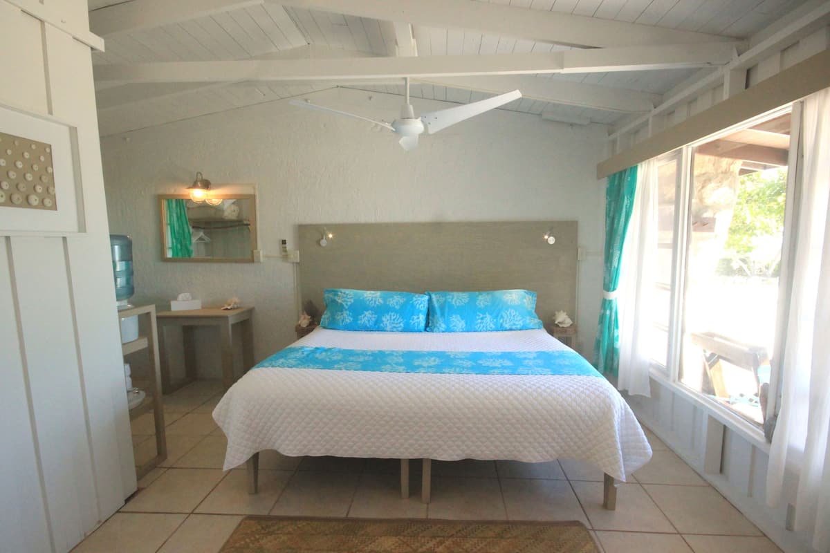 A bedroom with a bed, a ceiling fan, a window with curtains, and a small table with a lamp.
