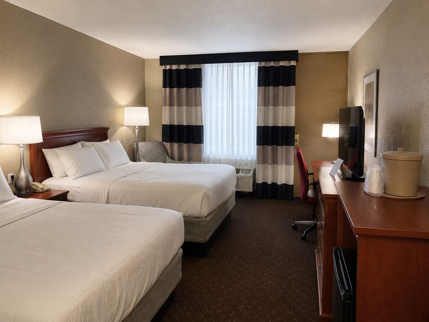 A hotel room with two beds, a desk with a TV, a chair, and a window with curtains.