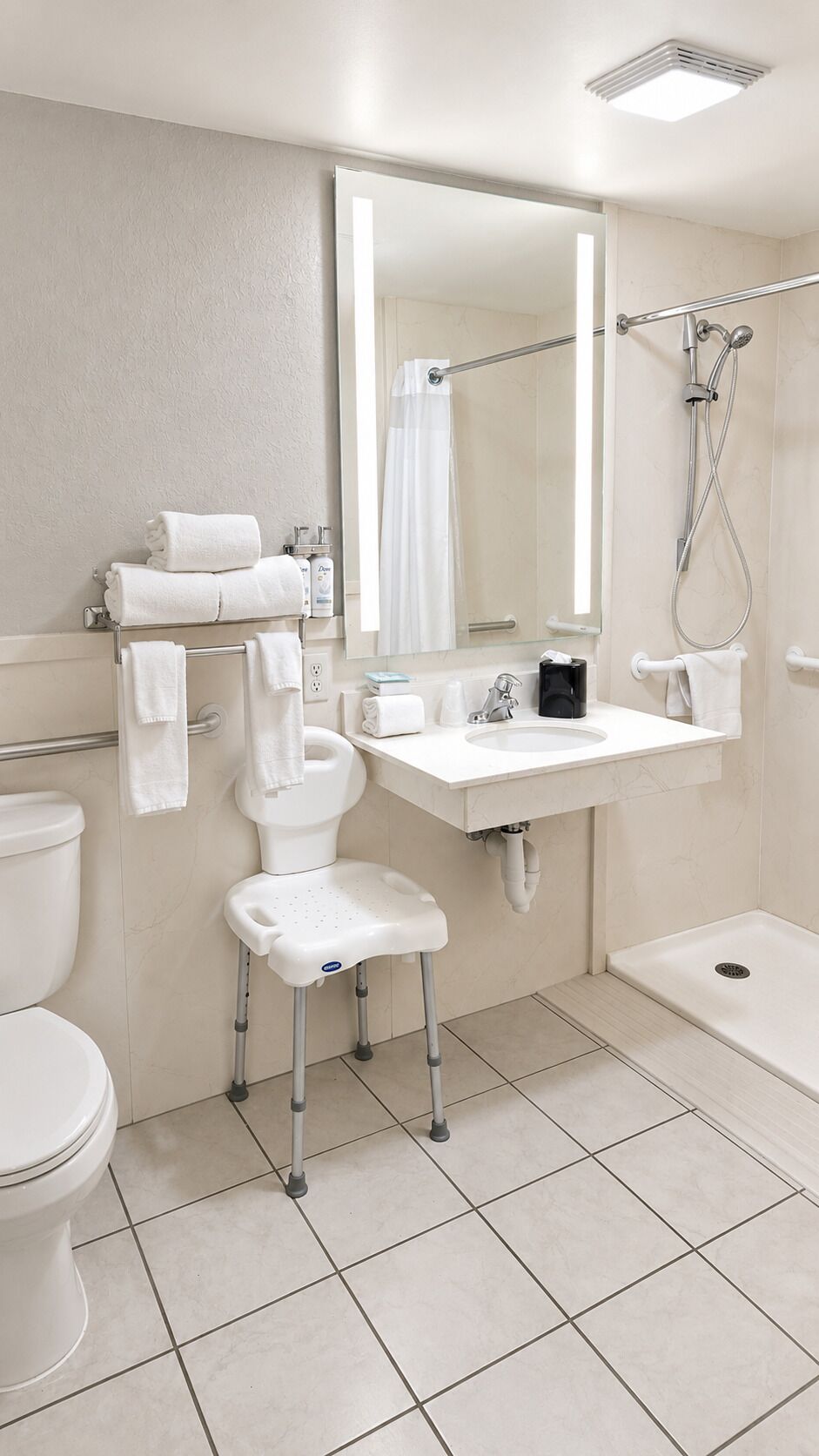 A bathroom with a toilet, a sink with a mirror, a shower, and a wheelchair-accessible chair.
