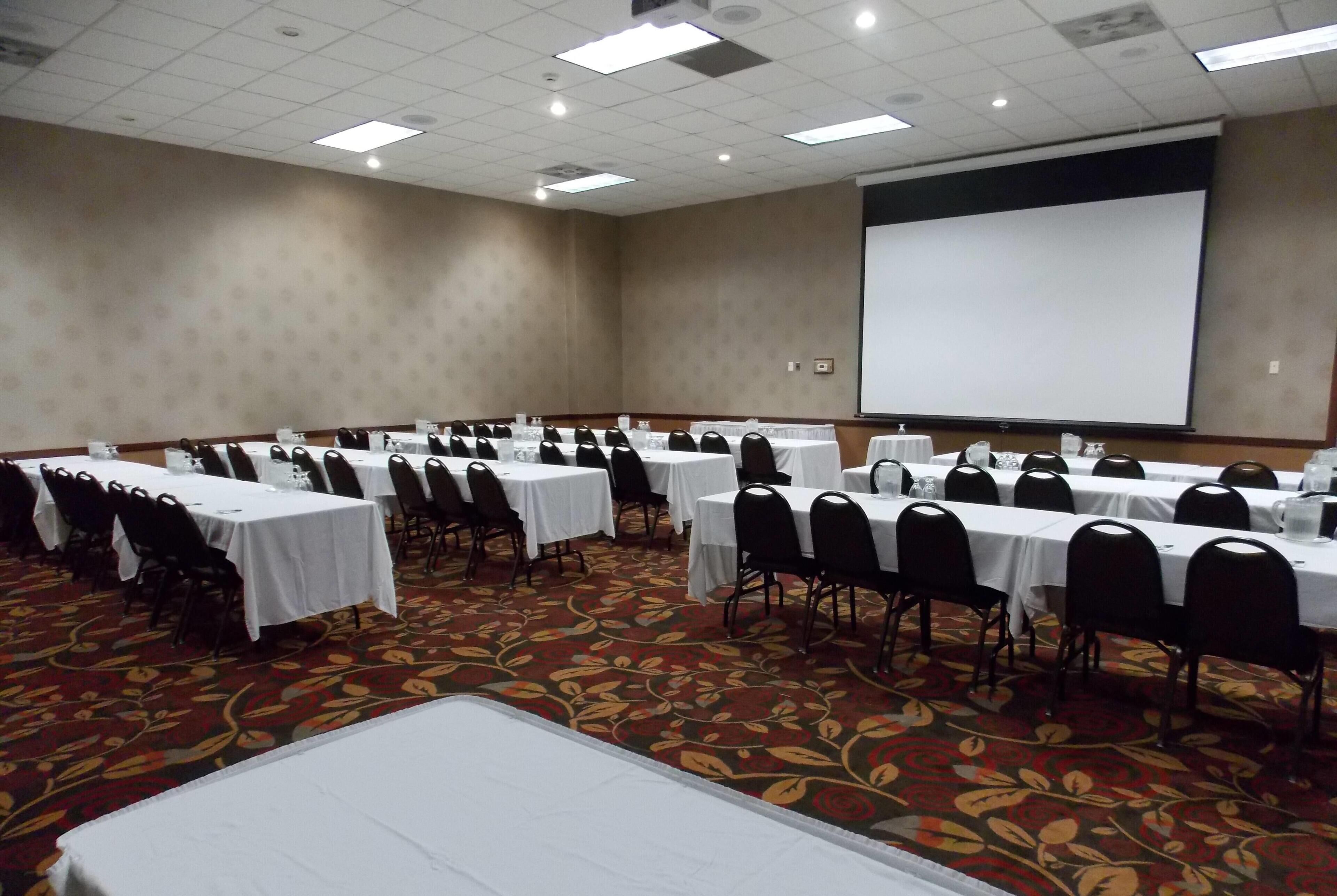 A conference room with multiple tables and chairs, a projector screen, and a carpeted floor.