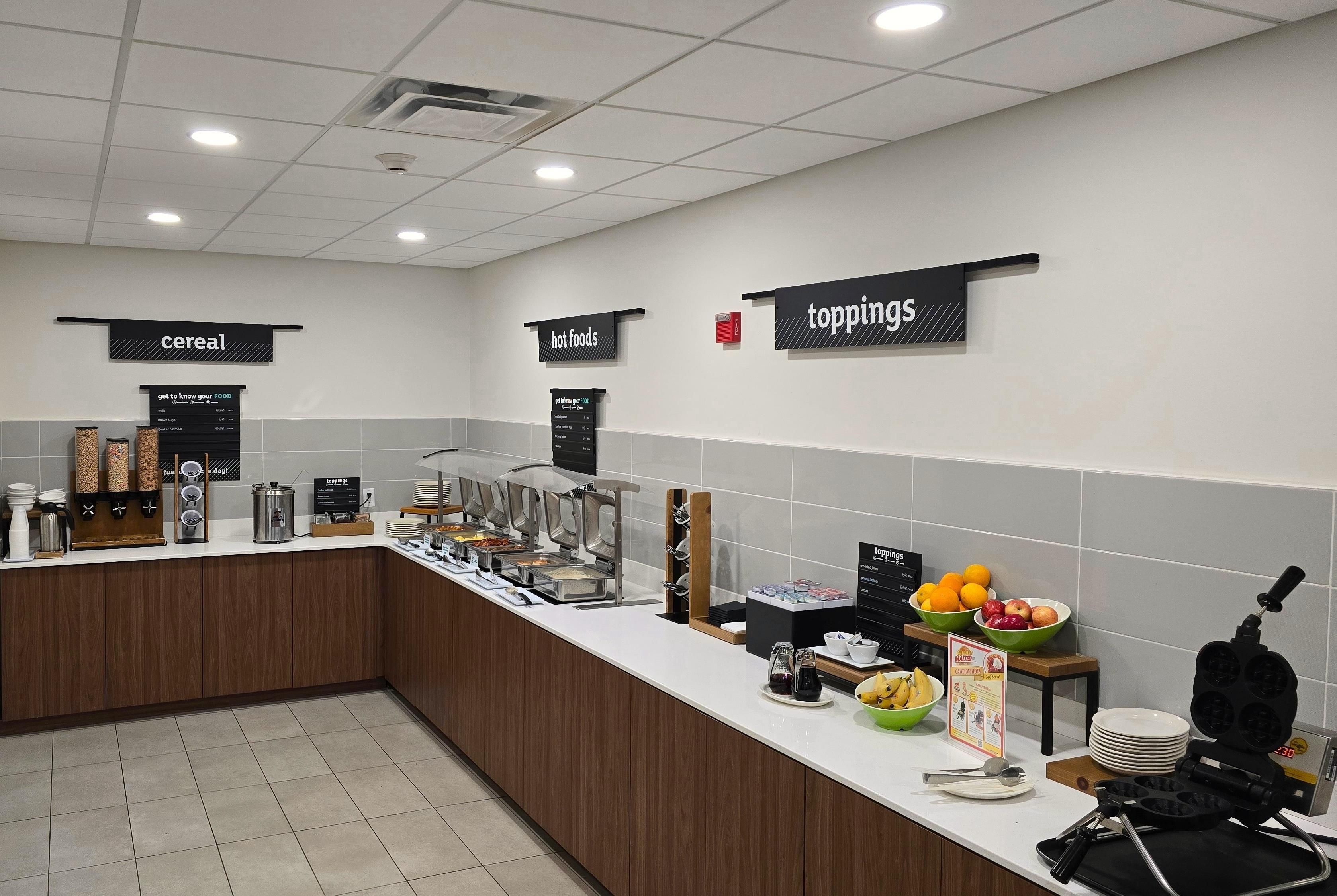 Zona de desayuno tipo buffet con secciones identificadas para cereales, alimentos calientes y aderezos.