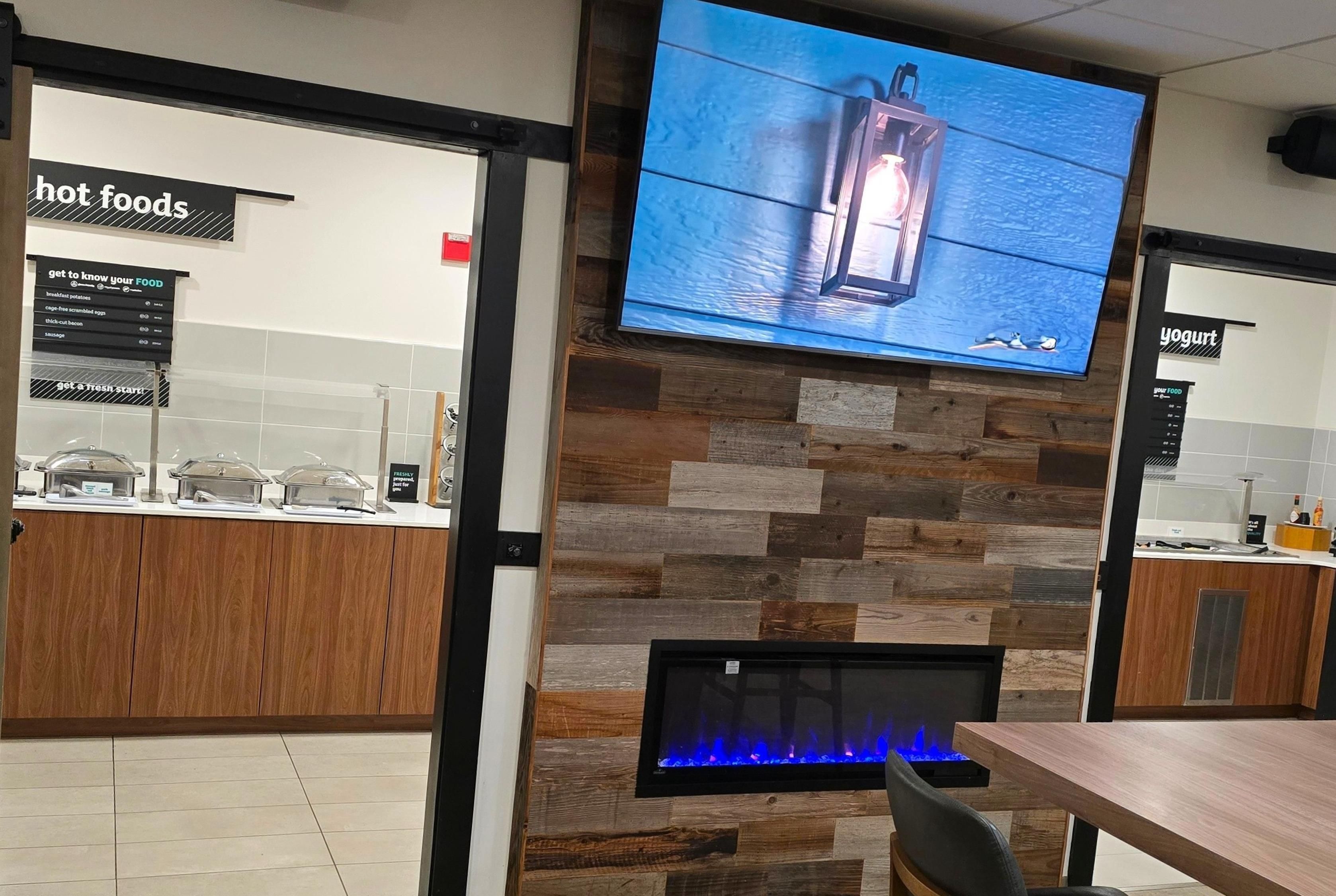 A restaurant interior with a wooden wall, a television mounted above a fireplace, and a counter displaying hot foods.