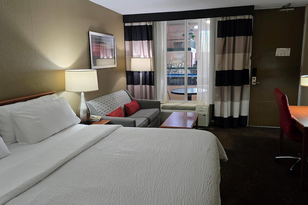 A hotel room with a bed, a sofa, a table, a lamp, a window with curtains, and a view of an outdoor balcony.