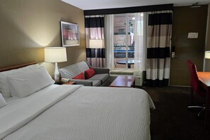 Hotellihuone, jossa on sänky, sohva, pöytä, lamppu, ikkuna verhoilla ja näkymä ulkoiselle parvekkeelle.