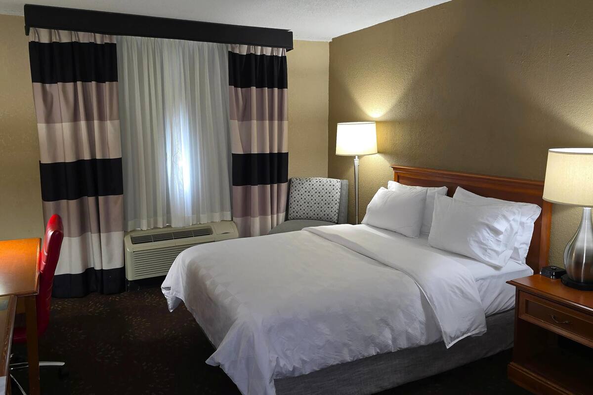 A hotel room with a bed, a desk, a chair, a lamp, and a window with curtains.