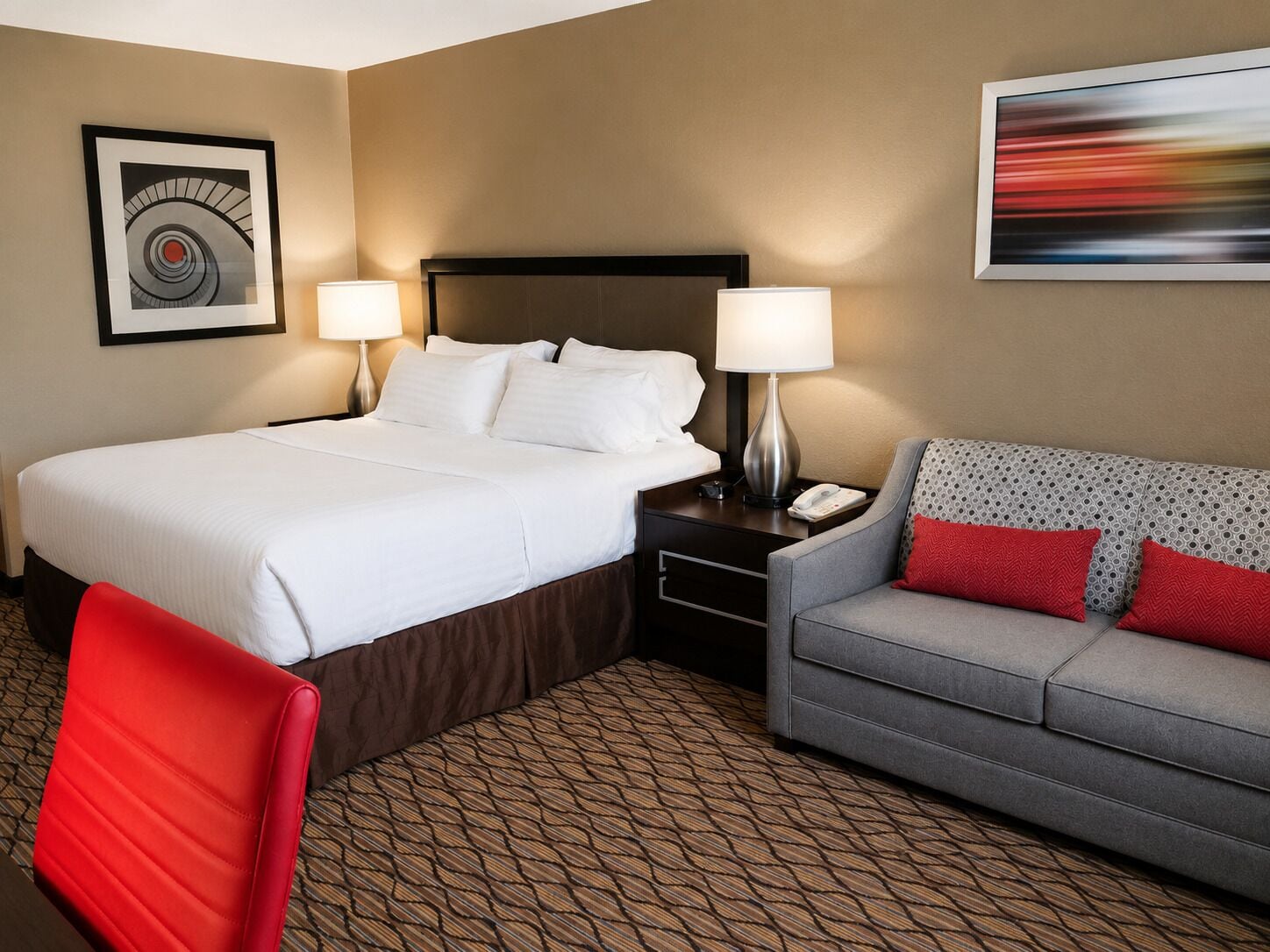 A hotel room with a large bed, two bedside tables with lamps, a grey sofa with red cushions, and patterned flooring.