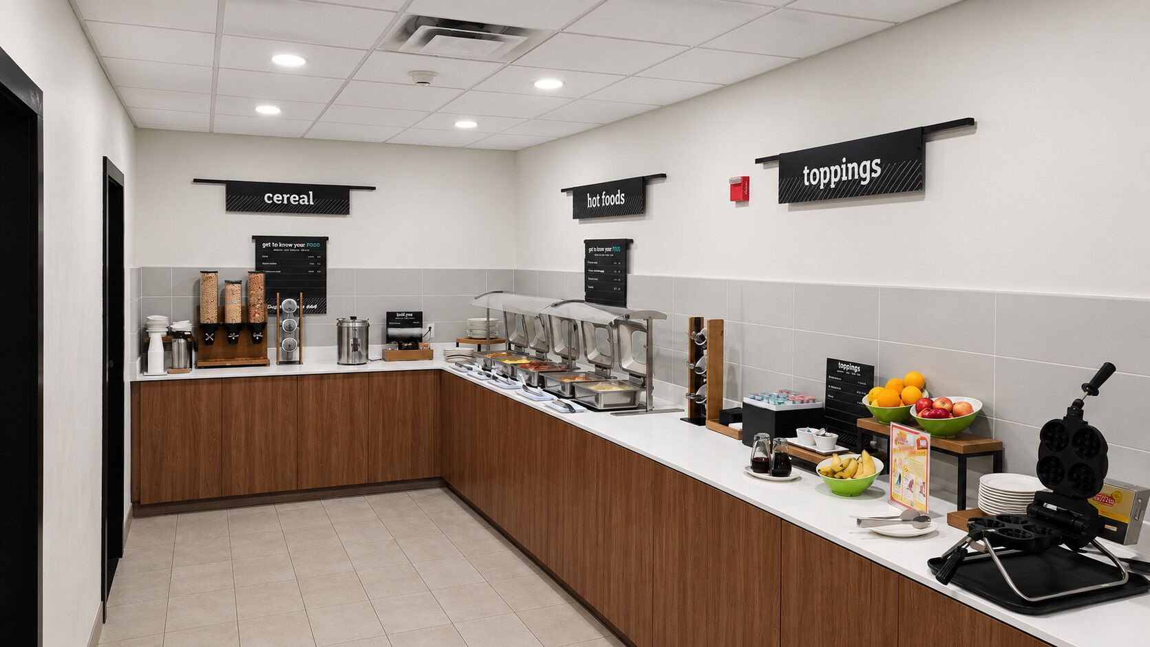 A modern breakfast bar with labeled sections for cereal, hot foods, and toppings, equipped with coffee machines and a waffle maker.