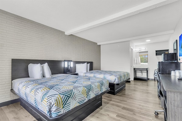 A modern hotel room with a brick accent wall, two beds with patterned bedding, a desk with a computer, and a view of the outdoors. - Motel 6 Summerville, SC – Charleston (Summerville)